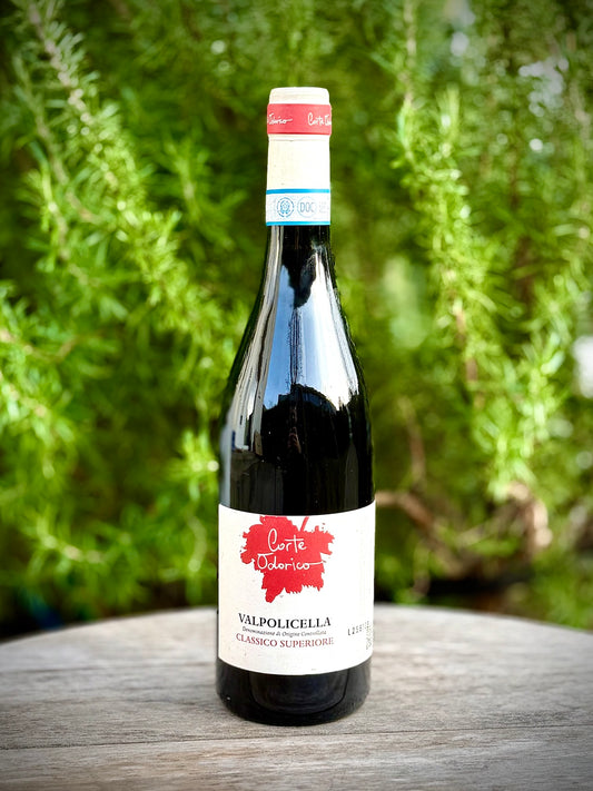 Eine Weinflasche vom Typ Valpolicella Classico Superiore von Corte Odorico mit einem roten Etikett, auf einer Holzoberfläche, mit grünem Pflanzenbewuchs im Hintergrund.