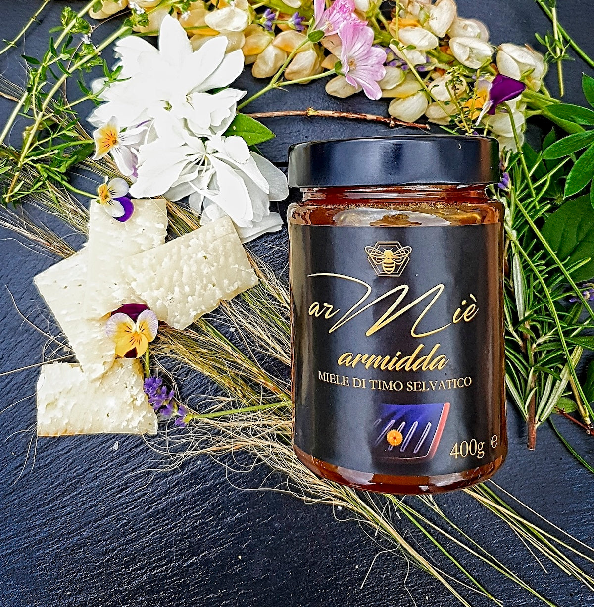 Glas mit sardischem Wildthymianhonig und einem dekorativen Blumenarrangement auf einer dunklen Oberfläche.