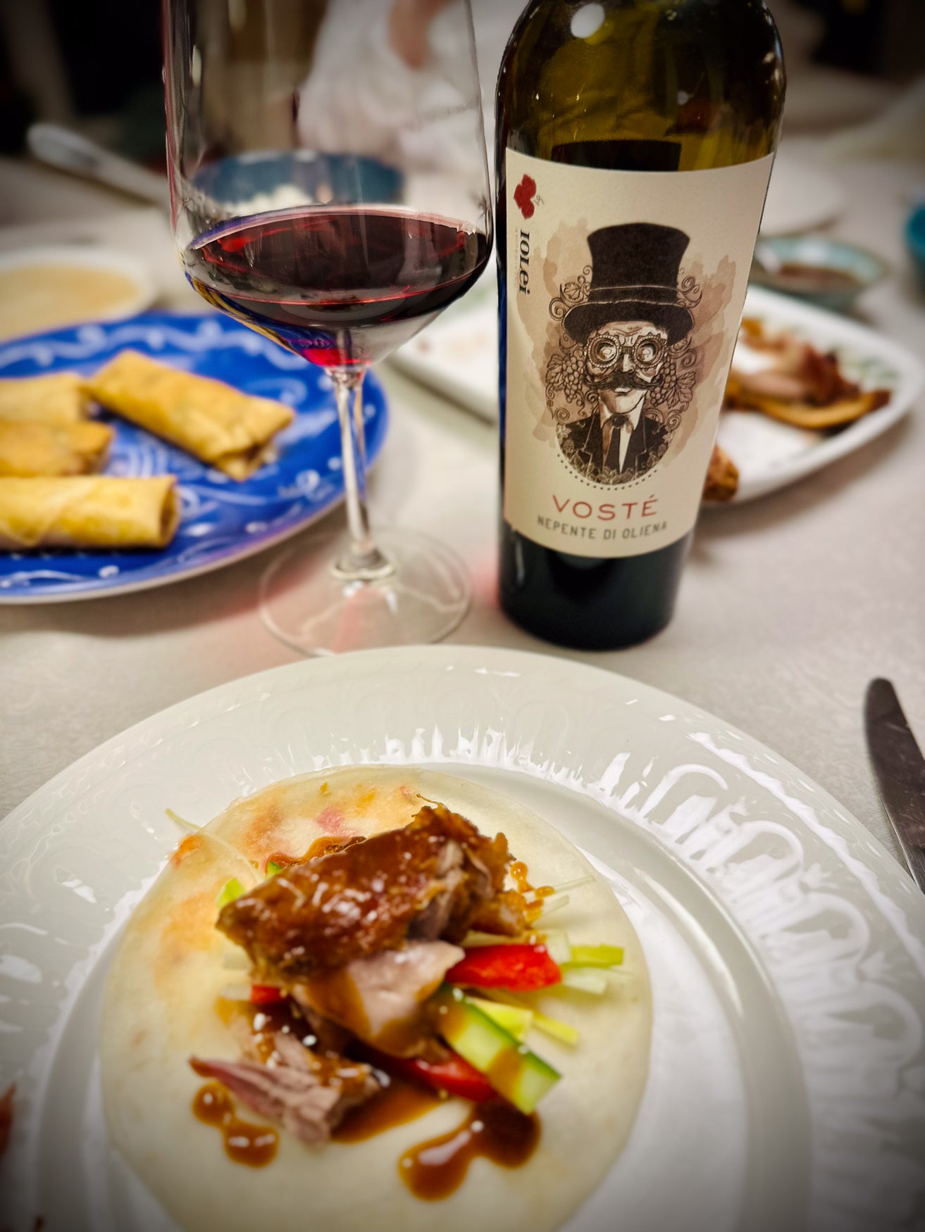 Dinner setting with a bottle of Voste wine, a glass of red wine, and a plate of food on a table.