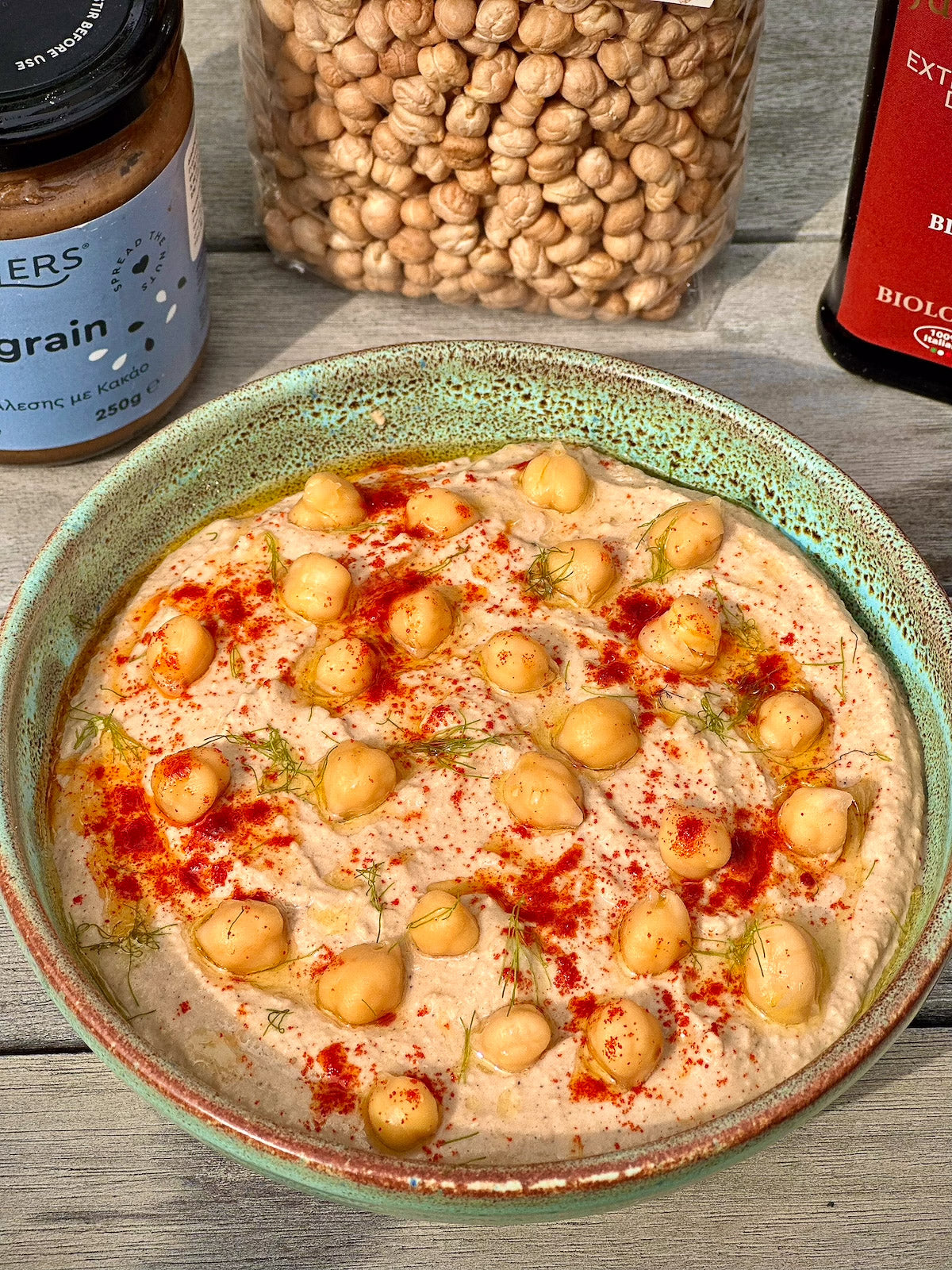 Hummus with chickpeas on a rustic wooden surface with jars of ingredients in the background.