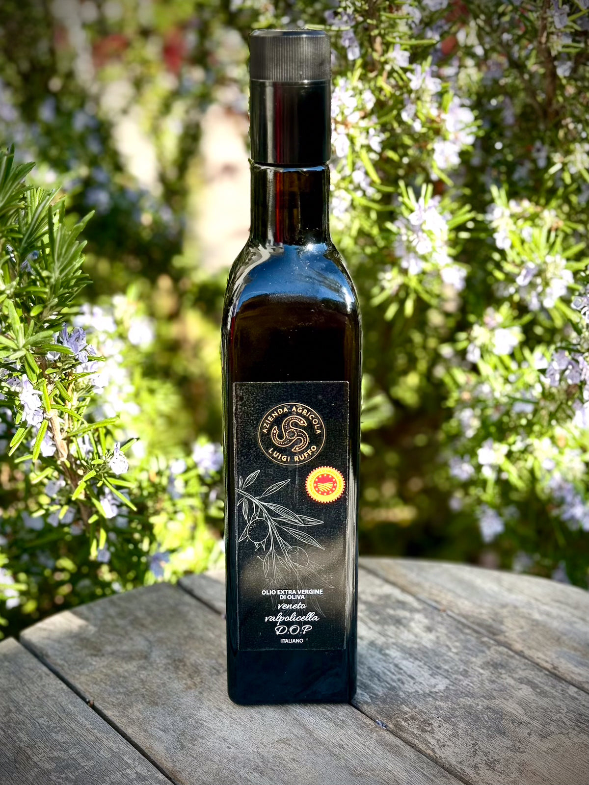 Bottle of olive oil with a label on a wooden surface with greenery in the background