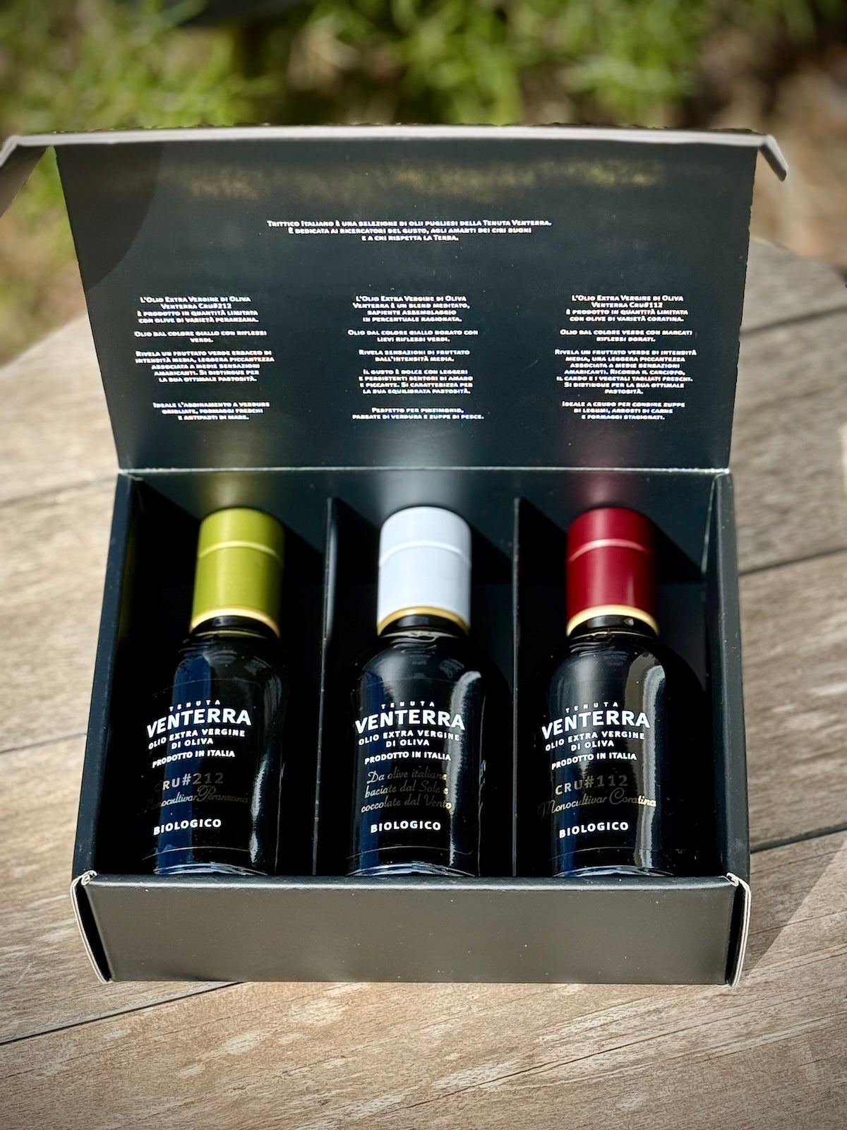 Three Venterra olive oil bottles in a box on a wooden surface with a blurred natural background