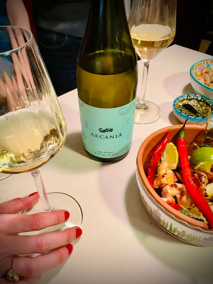 Wine bottle labeled 'Arcania' with two wine glasses and a dish of food on a table.