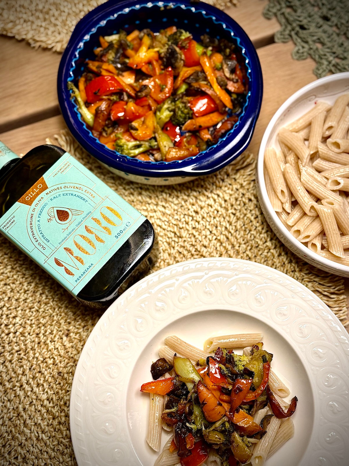 Dish of pasta with vegetables, bottle of Peranzana olive oil, and a bowl of pasta on a textured surface.