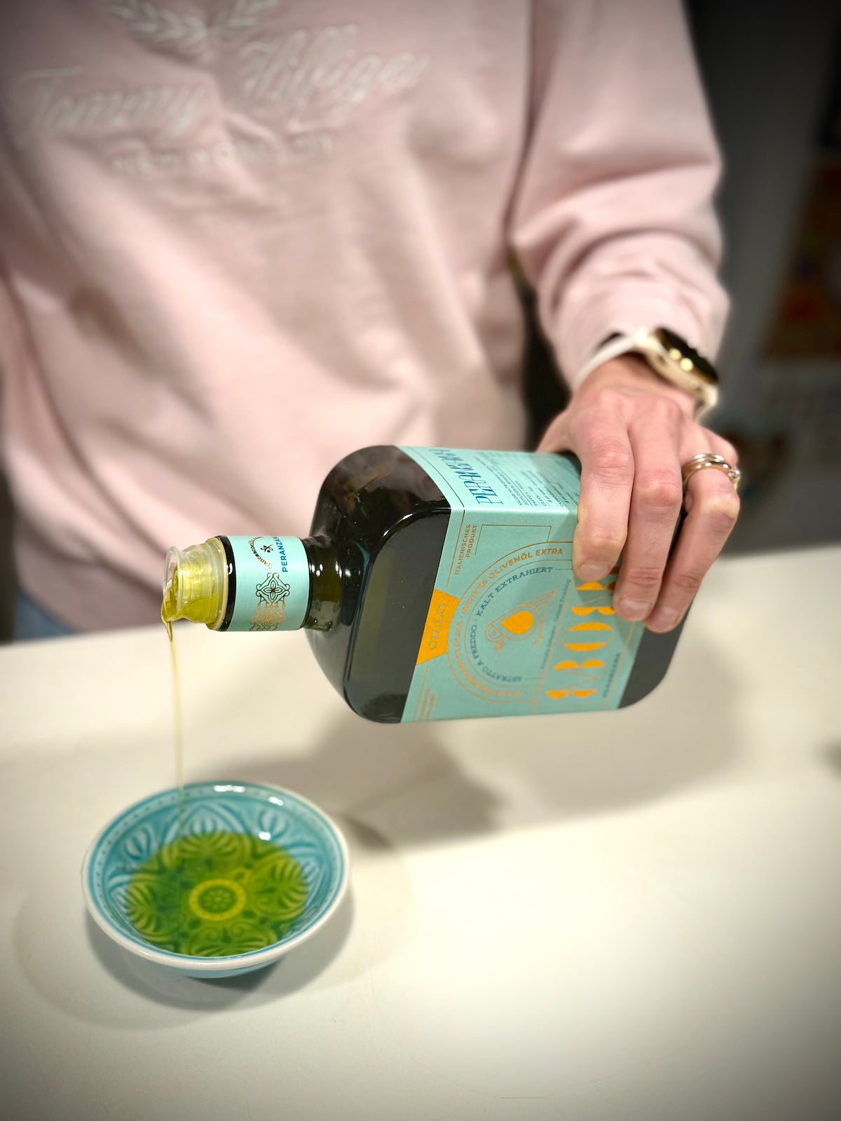 Person pouring oil from a bottle into a small bowl on a white surface.