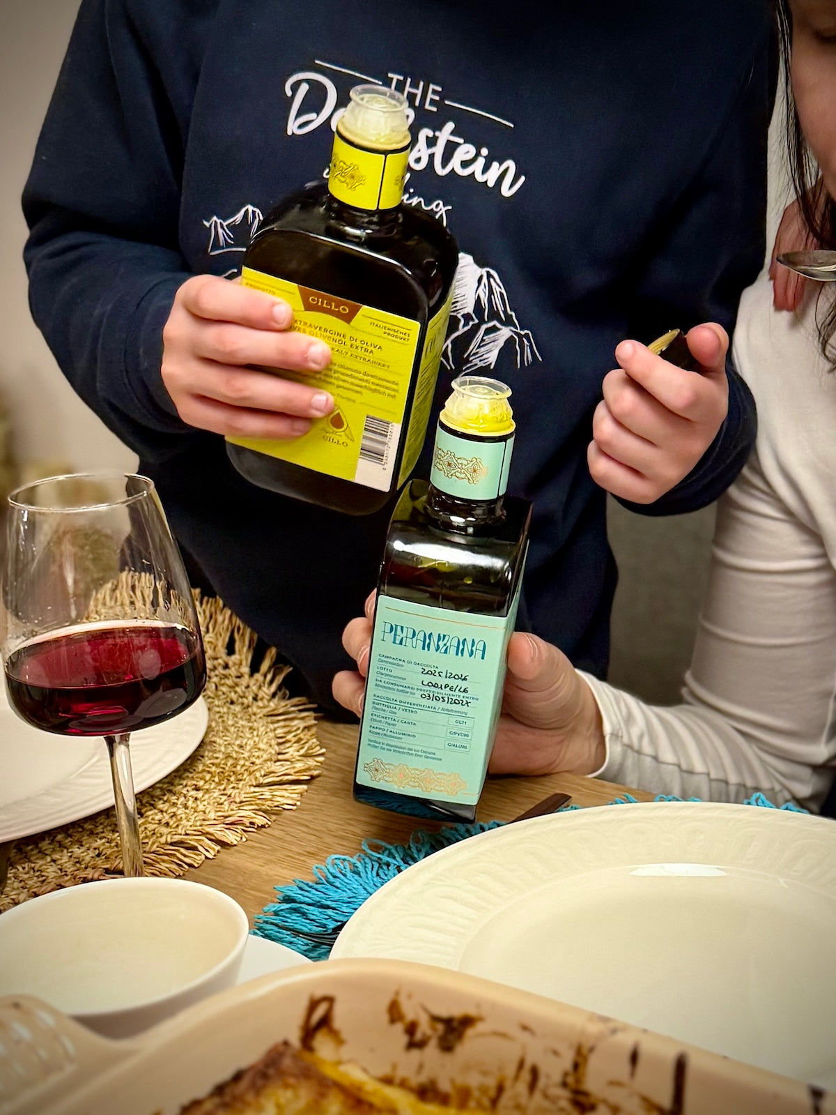 Two people holding bottles of Peranzana and Coratina olive oil at a table with a glass of red wine and food.