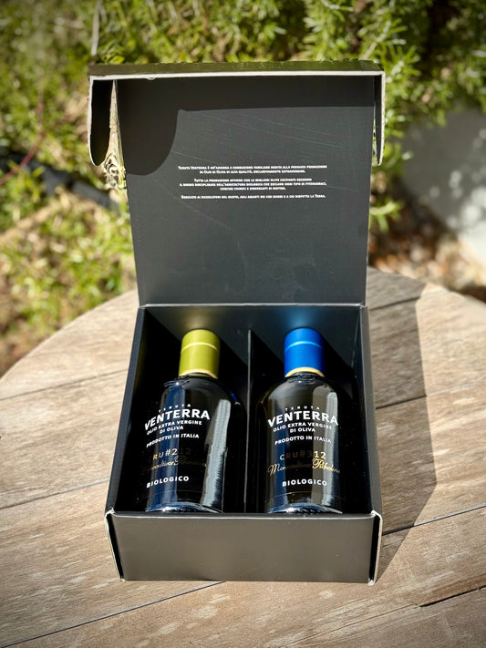 Two bottles of Venterra olive oil in a black box on a wooden surface with greenery in the background.