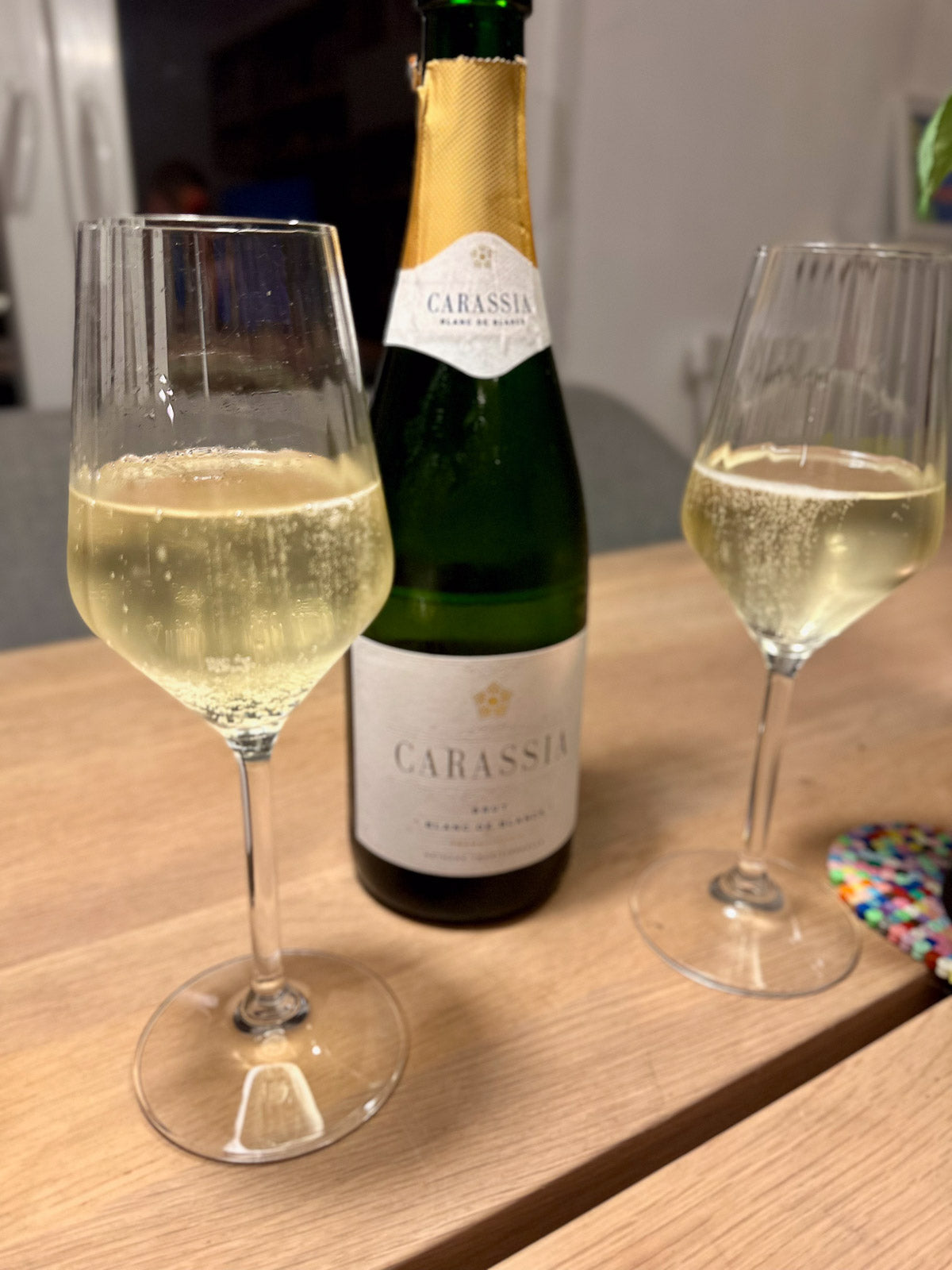 Two glasses of white wine and a bottle of Carassia blanc de blancs sparkling wine on a wooden table.