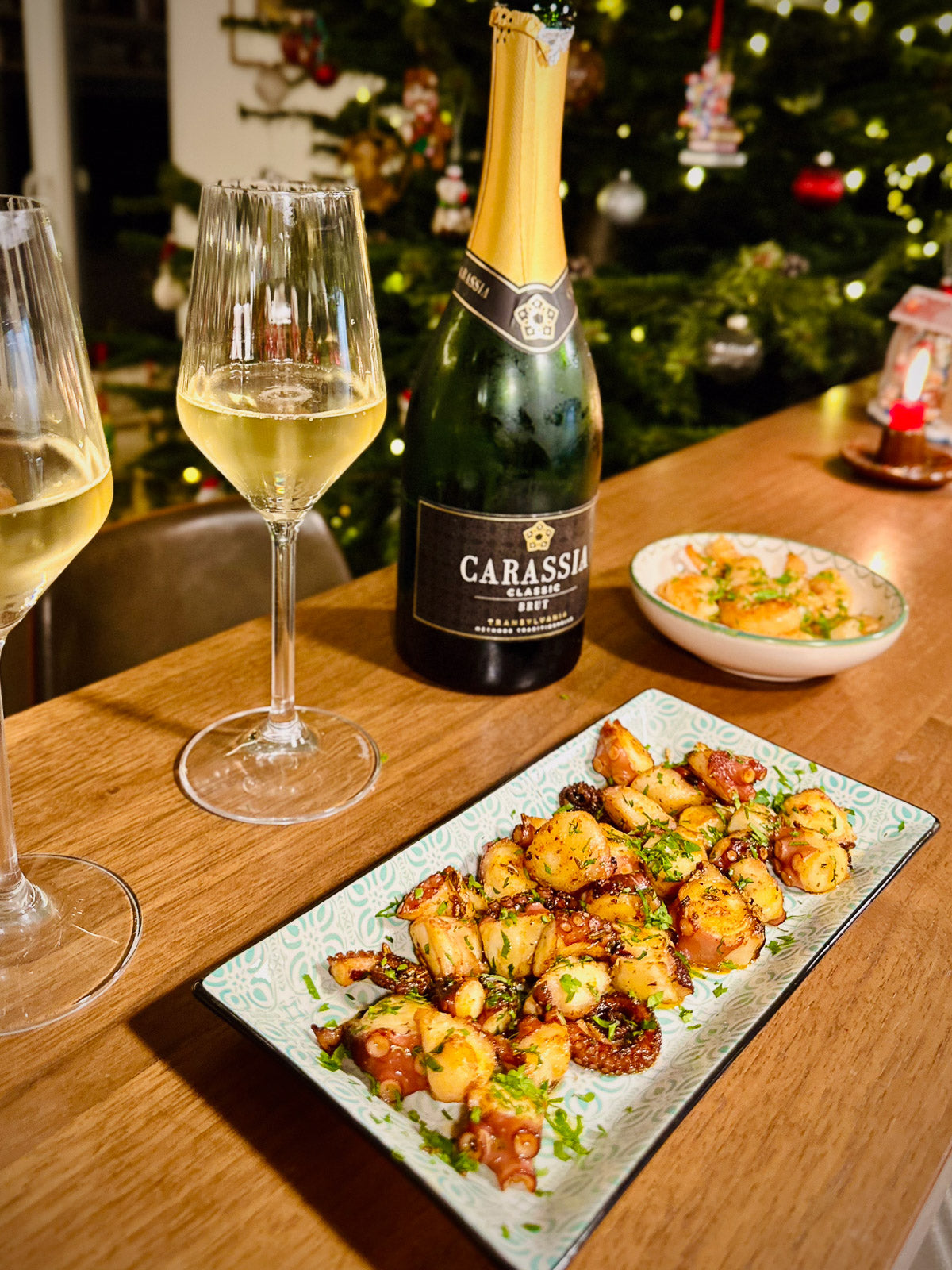 Bottle of Carassia wine with two glasses and a plate of food on a wooden table, Christmas tree in the background.
