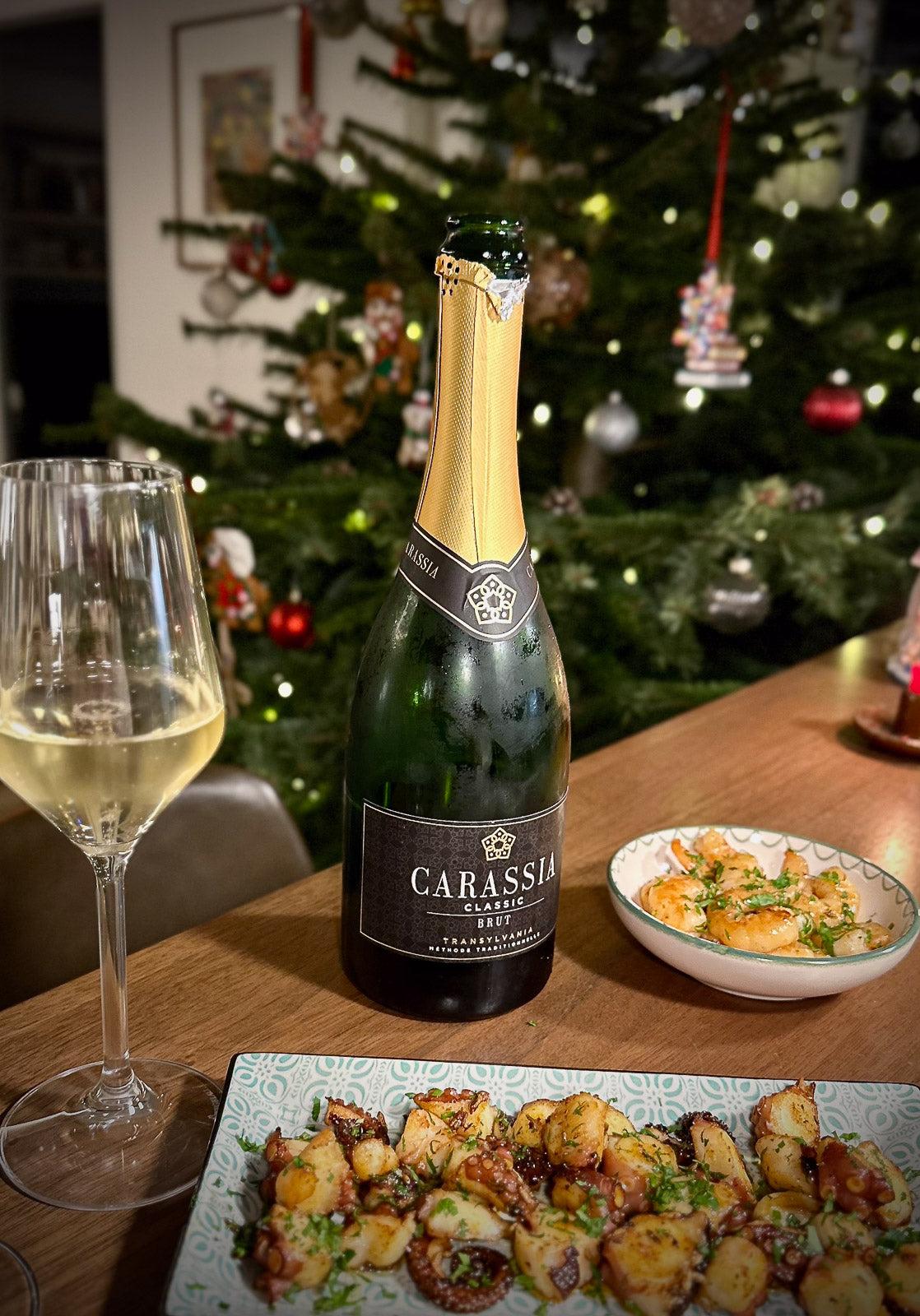 Bottle of Carassia brut with a glass and food on a table in front of a decorated Christmas tree.