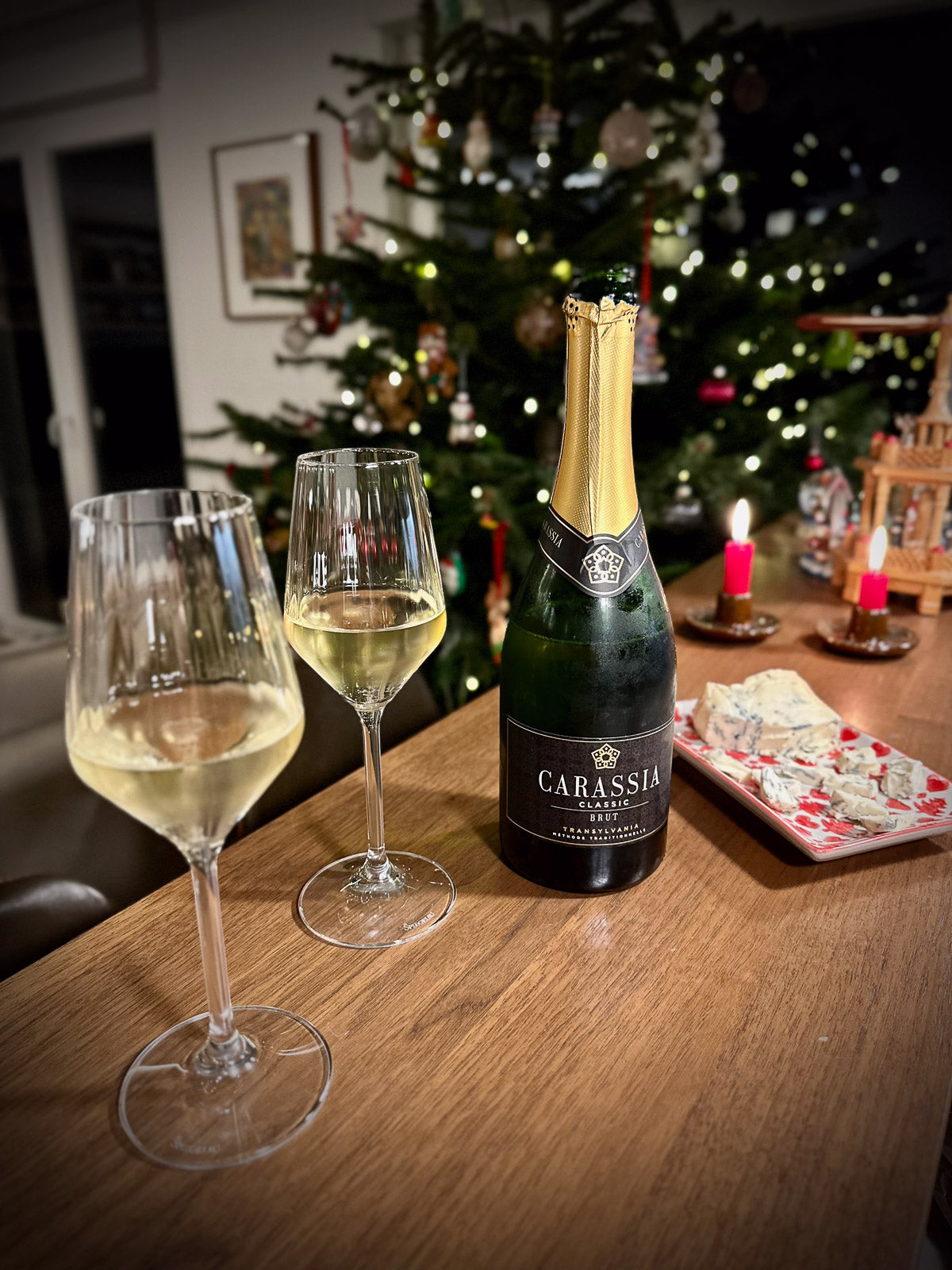 Bottle of Carassia sparkling wine with two glasses on a table in front of a decorated Christmas tree.