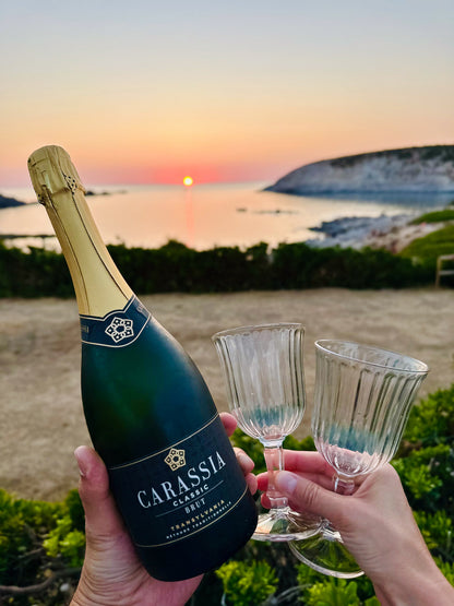 Bottle of Carassia sparkling wine with two glasses against a sunset beach backdrop
