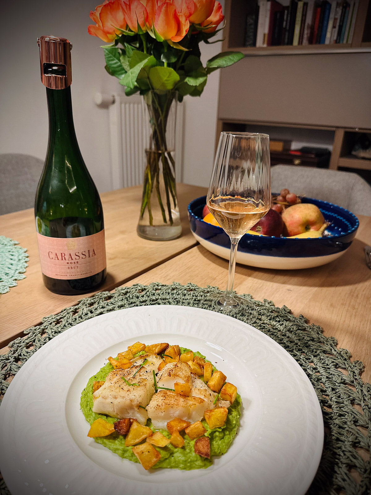 Dinner setting with a dish of fish and vegetables, a bottle of Carassia sparkling wine, and a glass on a table.