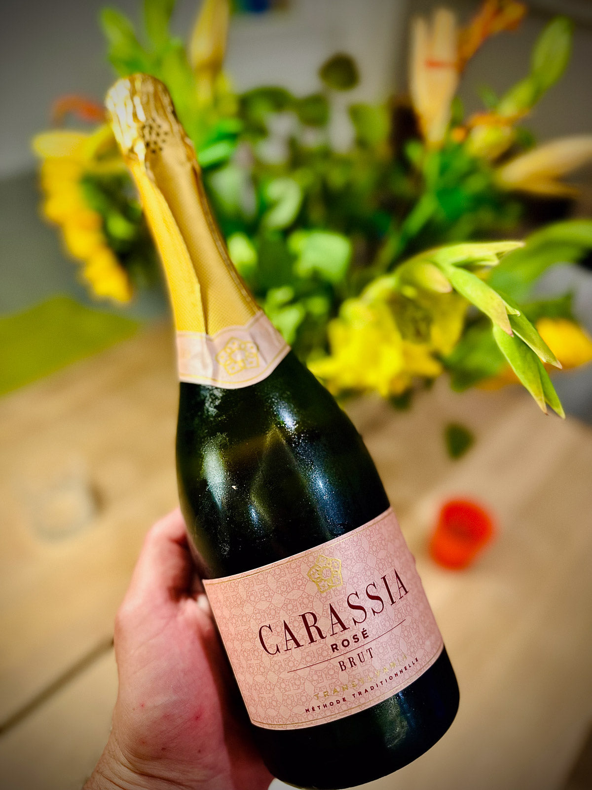 Bottle of Carassia Rose Brut sparkling wine held by a hand with flowers in the background