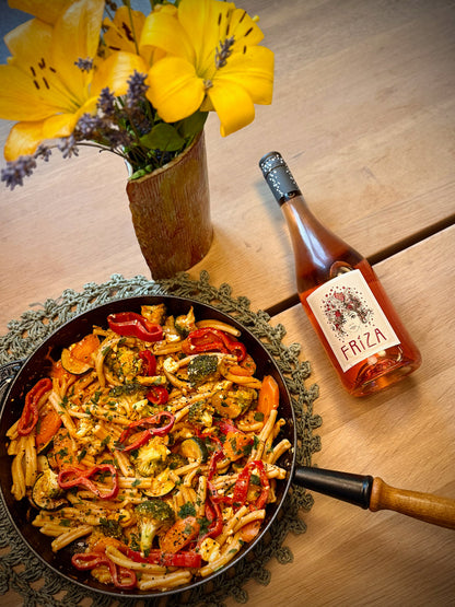 Fried pasta dish with vegetables next to a bottle of Friza on a wooden table with flowers.