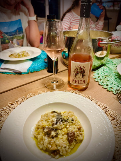 Table setting with a dish of risotto, a bottle of Friza wine, and a glass on a wooden table.