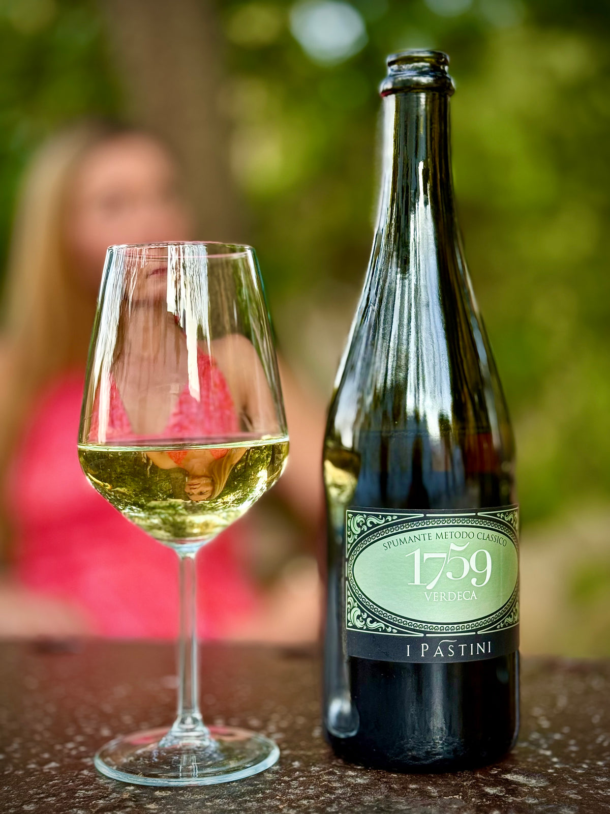 A bottle of 1759 Pastini Verdeca sparkling wine next to a filled wine glass on a stone surface, with a blurred person in a pink top in the background outdoors.