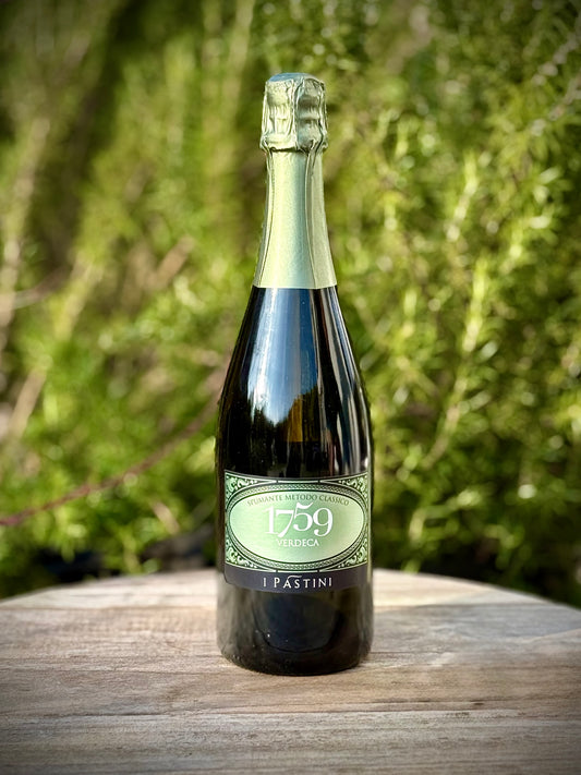 A bottle of 1759 Pastini Verdeca sparkling wine, standing on a rustic wooden barrel with a blurred green garden background.