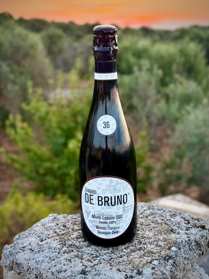A bottle of Sandro De Bruno sparkling wine with a dark glass body and white label, standing on a stone surface with a blurred outdoor landscape in the background.