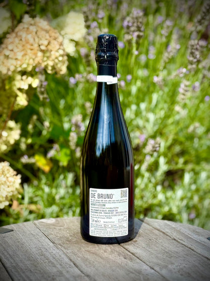 A bottle of Sandro De Bruno Monti Lessini sparkling wine stands on a wooden table outdoors, with a blurred garden background of green foliage and white flowers.