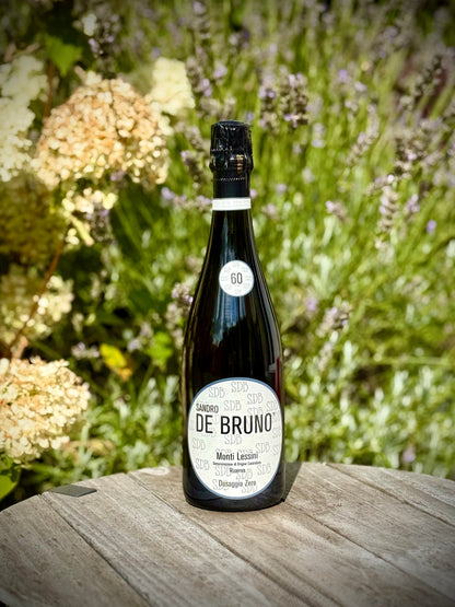 A bottle of Sandro De Bruno Monti Lessini sparkling wine stands on a wooden table outdoors, with a blurred garden background of green foliage and white flowers.
