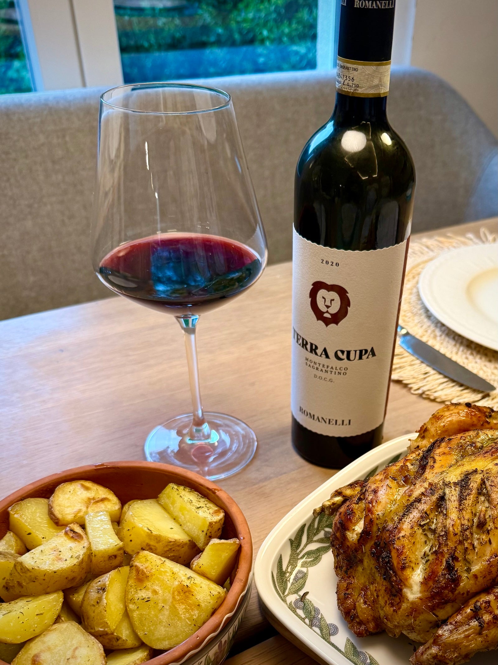 Bottle of Terra Cupa red wine with a glass, roasted potatoes, and roasted chicken on a table.